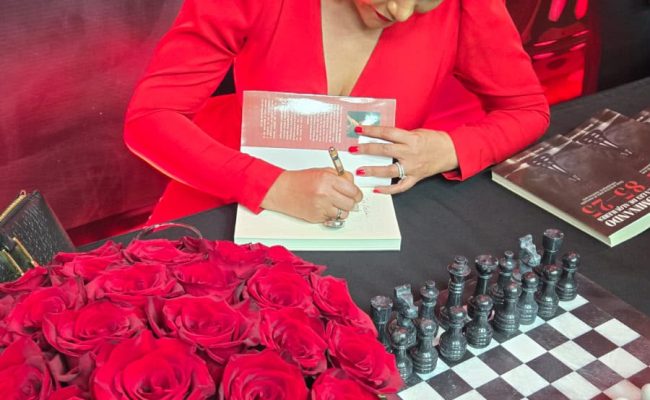 Mujer firmando libro junto a rosas y ajedrez